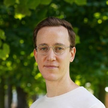 A portrait of Gabe standing in the courtyard of Designmuseum in Copenhagen. Gabe is wearing a white t-shirt and wireframe glasses and is smiling. The green leaves of trees are out of focus in the background.