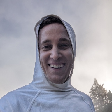 A portrait of Gabe, smiling and wearing a light hooded jacket with the hood up, outdoors under a cloudy sky.