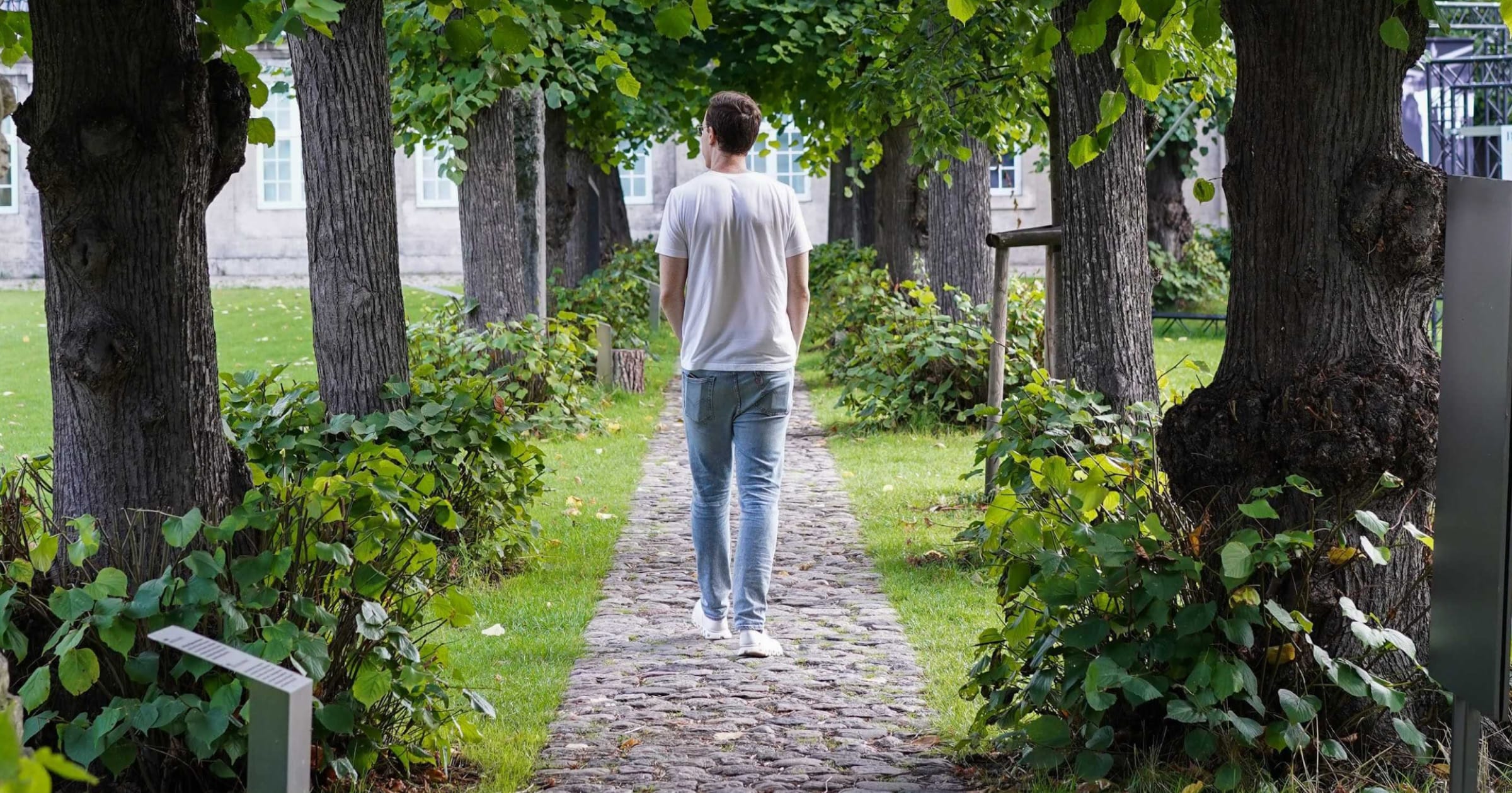 A photo of Gabe walking down a stone path in the courtyard of Designmuseum in Copenhagen. Gabe has his back turned to camera is walking away. He is wearing a white t-shirt, blue jeans, and white athletic shoes.